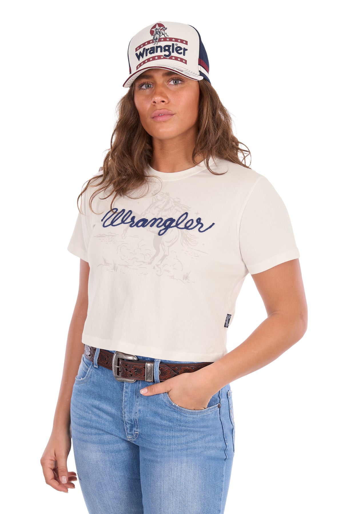 A woman wearing a white Wrangler short-sleeve crop top t-shirt with a navy blue script logo, paired with blue jeans and a white Wrangler trucker hat, against a plain white background.