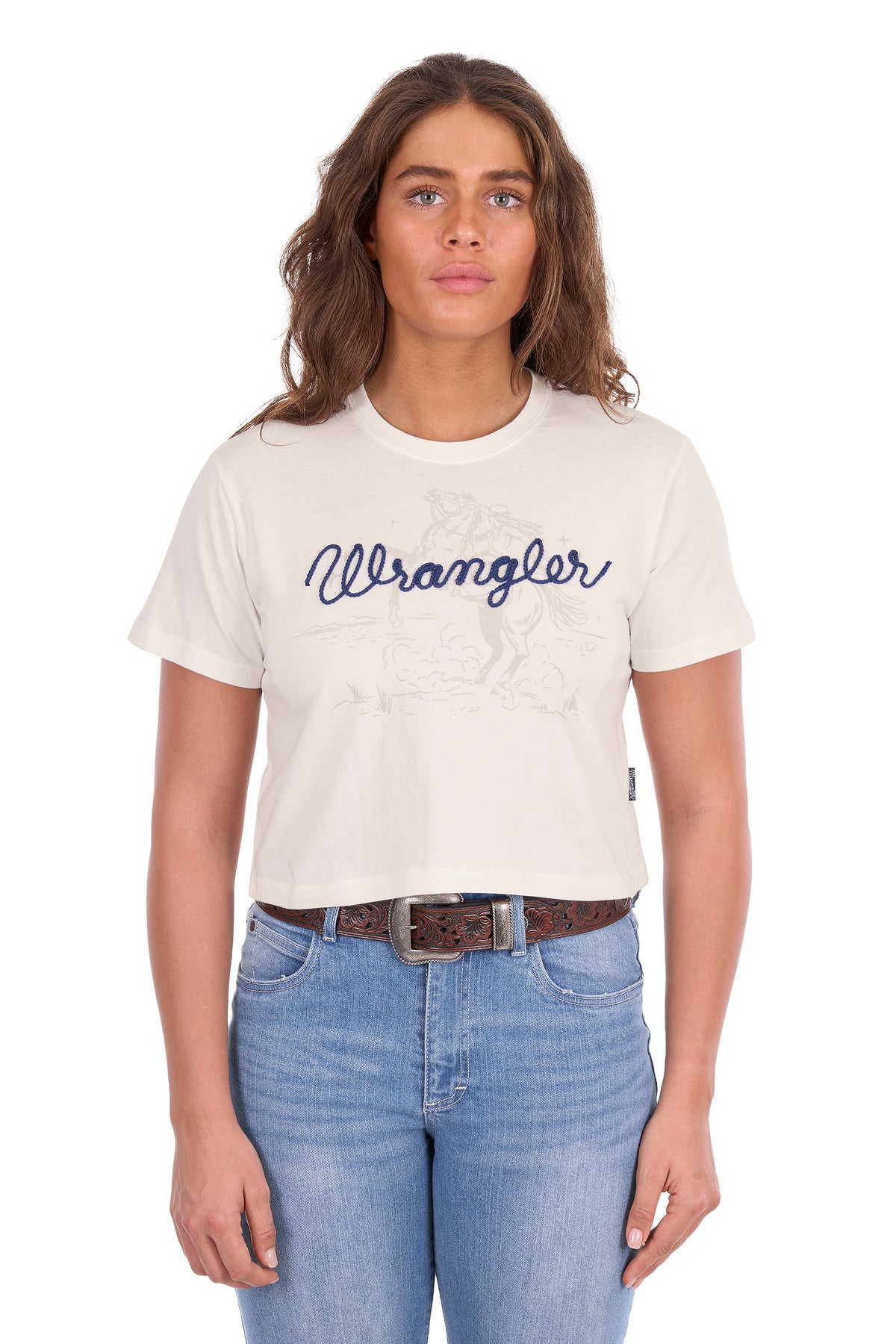 A woman modeling a short-sleeve crewneck t-shirt in off-white with a navy blue 'Wrangler' script logo across the chest, paired with blue jeans and a brown belt, against a plain white background.