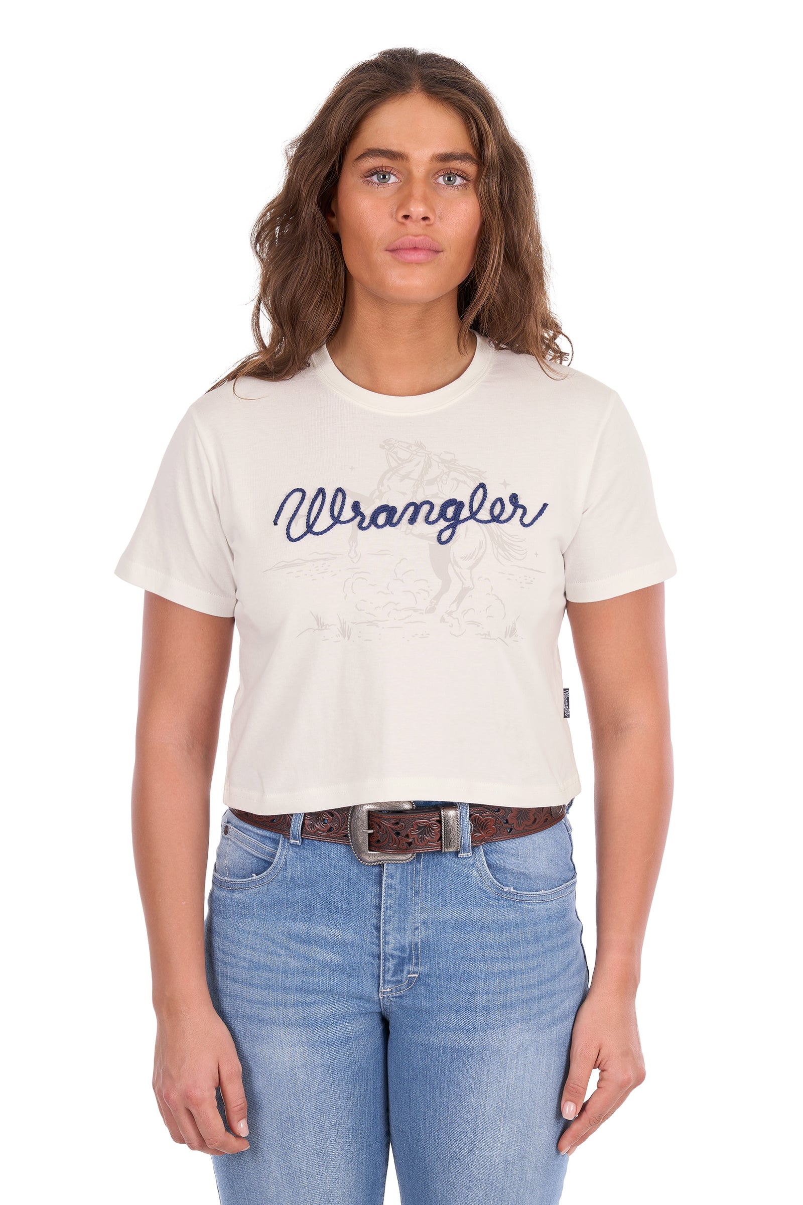 A woman modeling a short-sleeve crewneck t-shirt in off-white with a navy blue 'Wrangler' script logo across the chest, paired with blue jeans and a brown belt, against a plain white background.