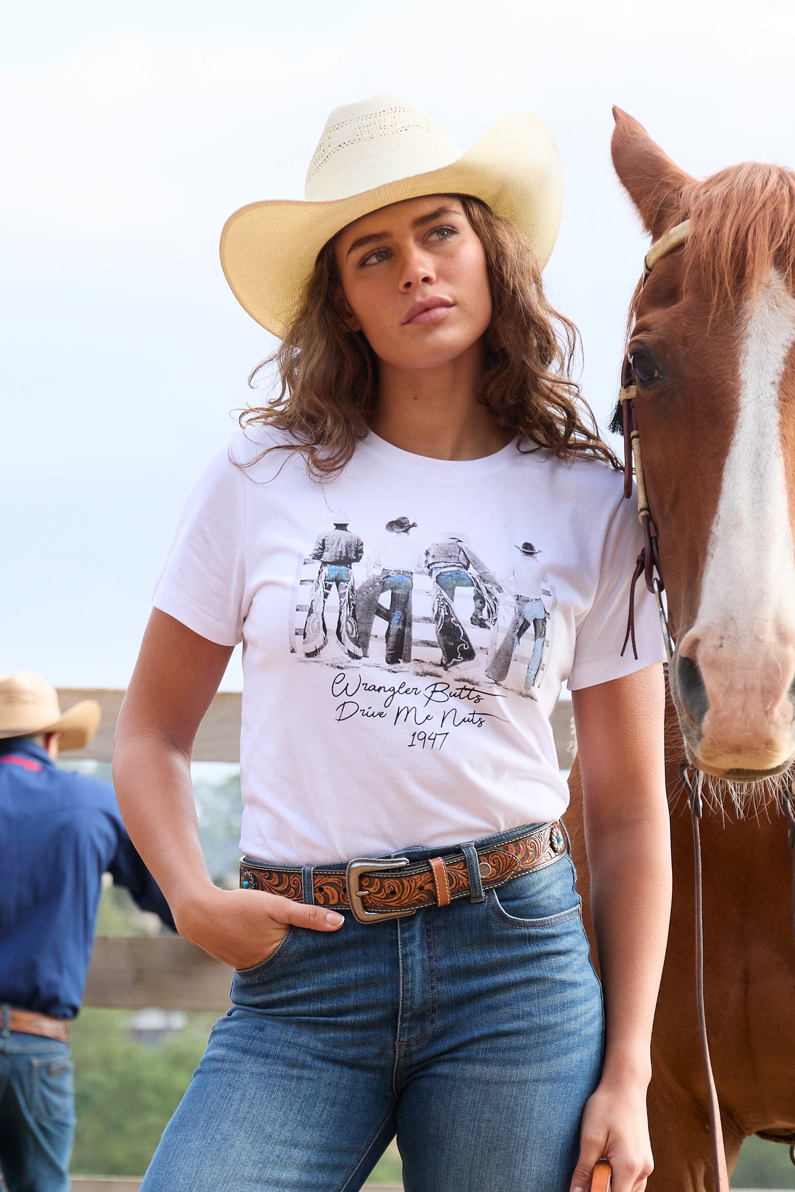 A woman wearing a white graphic t-shirt with a cowboy and cattle print, blue jeans, and a cream cowboy hat, standing next to a brown horse outdoors.