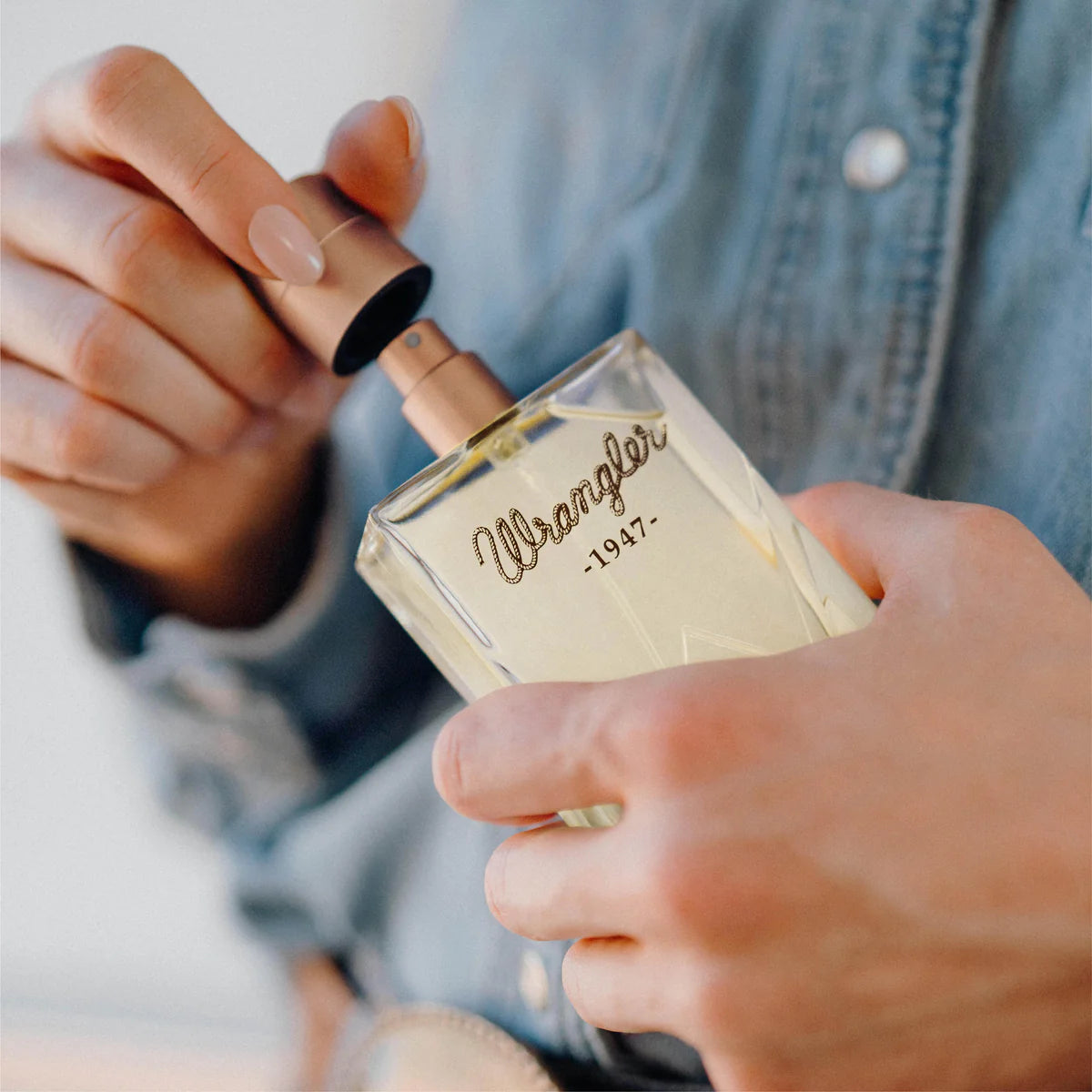 Wrangler Women's 1947 Eau De Parfum. Person holding a 'Wrangler 1947' perfume bottle with a denim jacket.