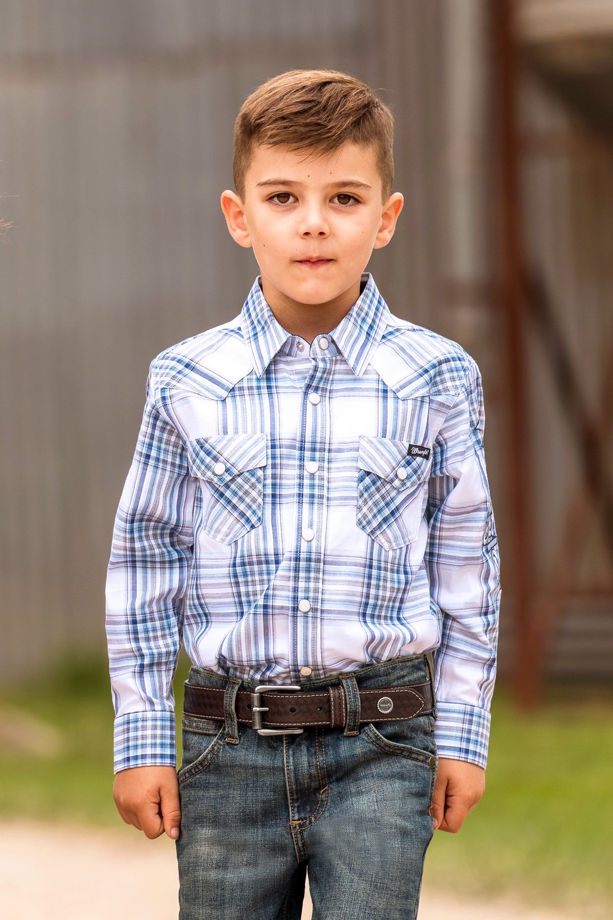 A young boy wearing a blue and white plaid long-sleeve western shirt with blue jeans and a brown belt, standing outdoors against a rustic wooden background.