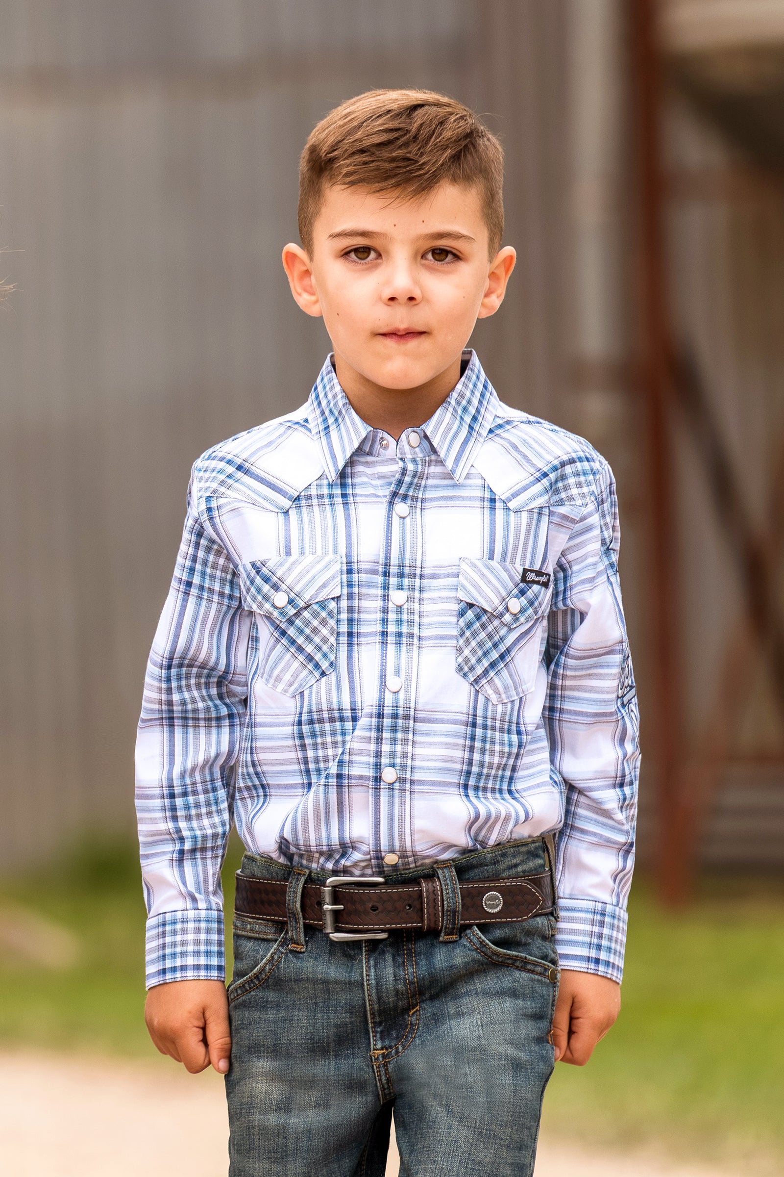 A young boy wearing a blue and white plaid long-sleeve western shirt with blue jeans and a brown belt, standing outdoors against a rustic wooden background.