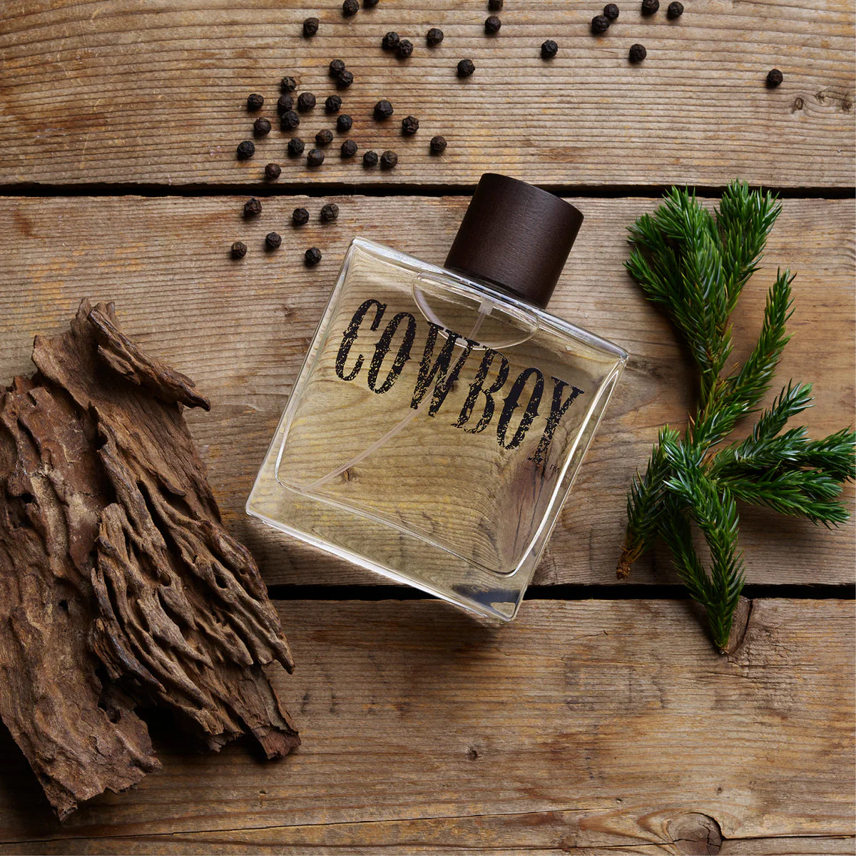 A clear rectangular perfume bottle labeled 'COWBOY' with a dark brown cap, placed on a rustic wooden surface with scattered black peppercorns, dried bark, and a small green pine sprig.