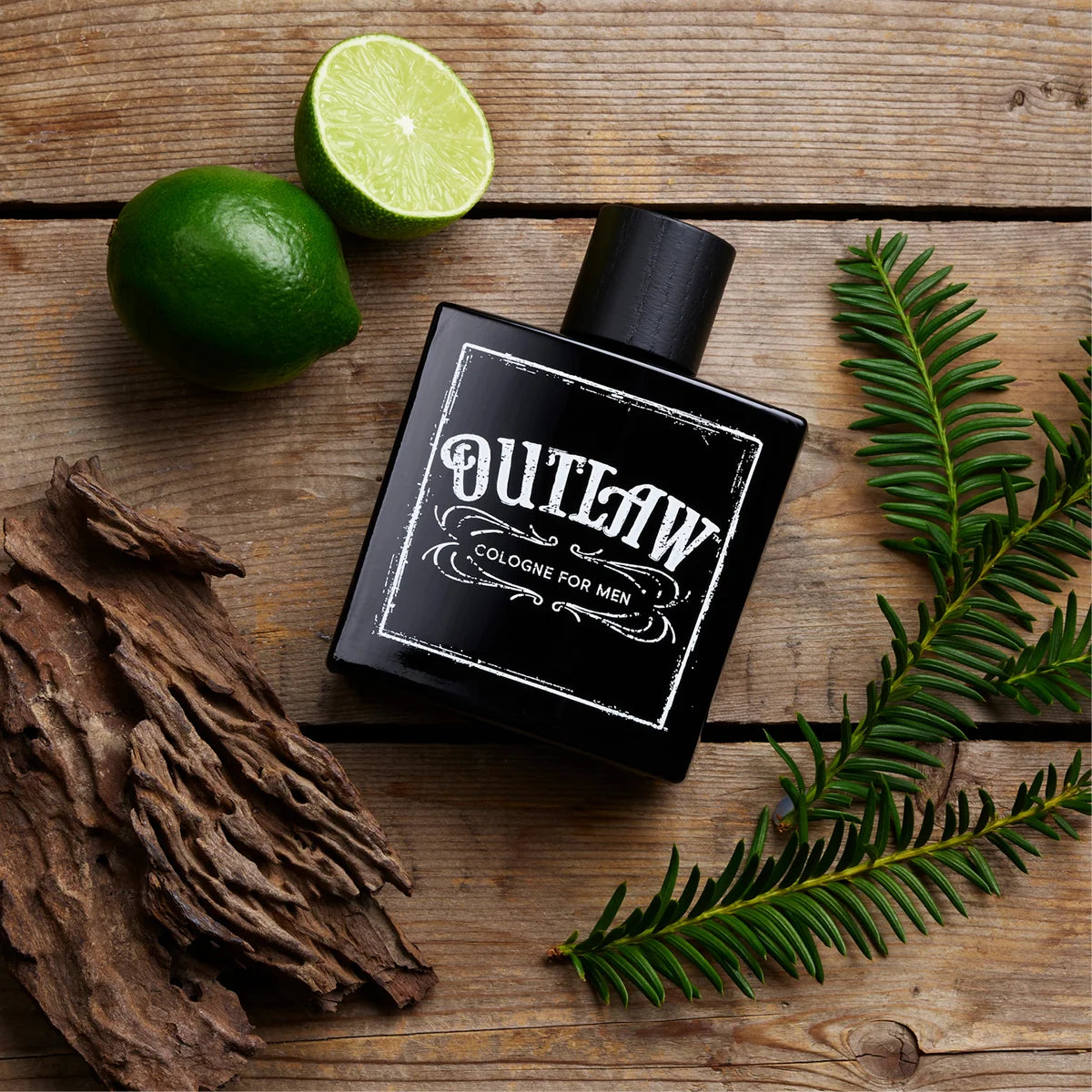 A black rectangular bottle of Outlaw Cologne for Men with a white distressed label, styled on a rustic wooden surface with limes, pine needles, and dried bark.