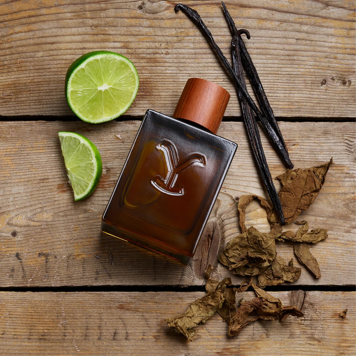 Perfume bottle with lime slices, vanilla beans, and dried leaves on a wooden surface