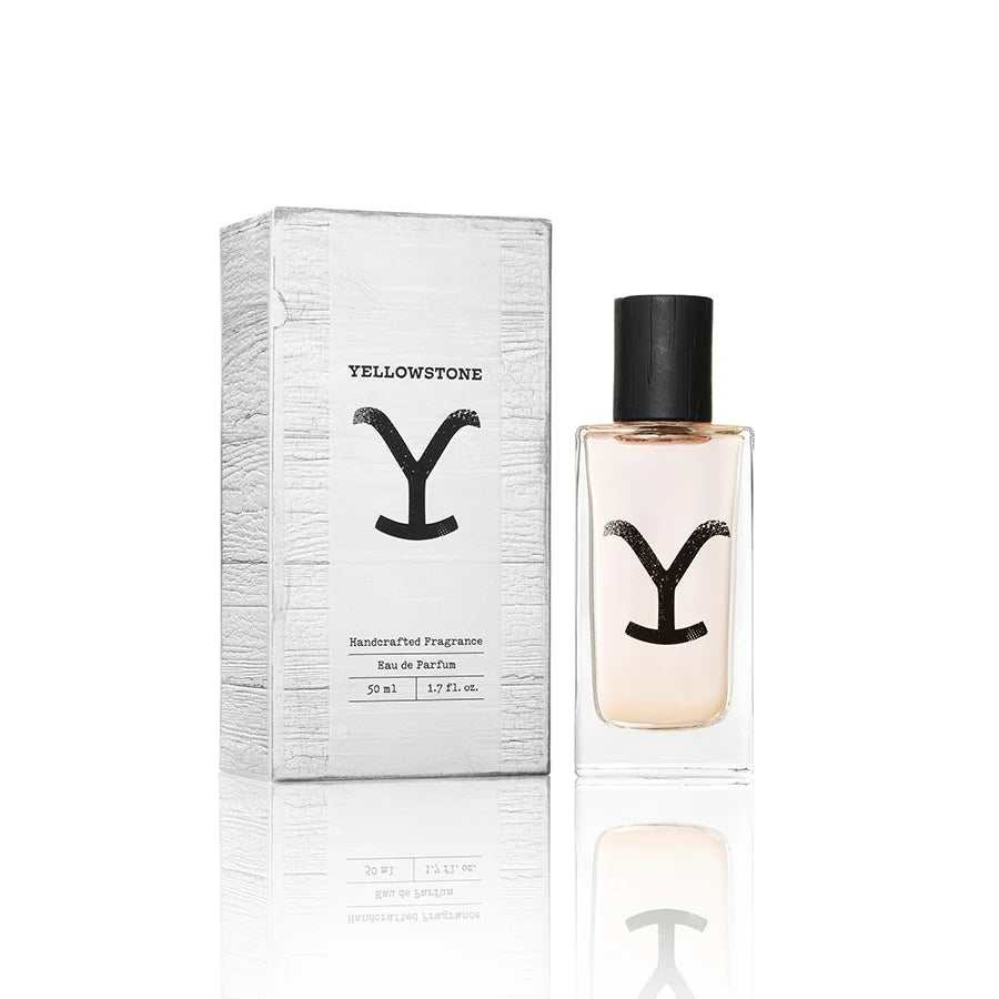 A clear rectangular glass perfume bottle with a black cap and a black stylized 'Y' logo on the front, containing a pale peach-pink liquid, shown on a white background with a soft reflection below.