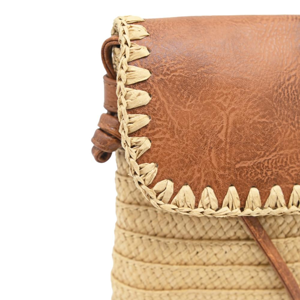 Close-up of a woven straw and leather bag corner showing a brown textured leather flap with decorative braided trim and a natural straw body on a white background.