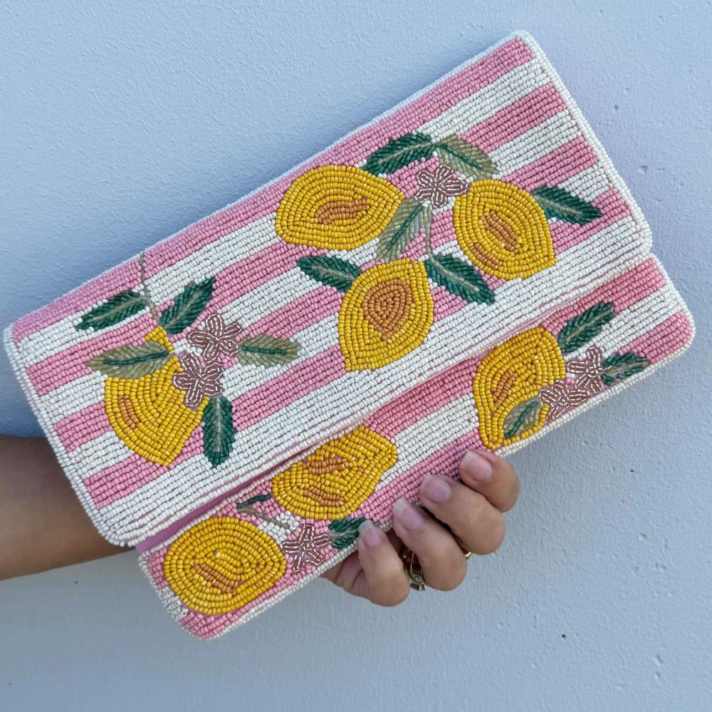 A hand holding a rectangular beaded clutch bag with a pink and white striped pattern and yellow lemon motifs, set against a light blue wall.