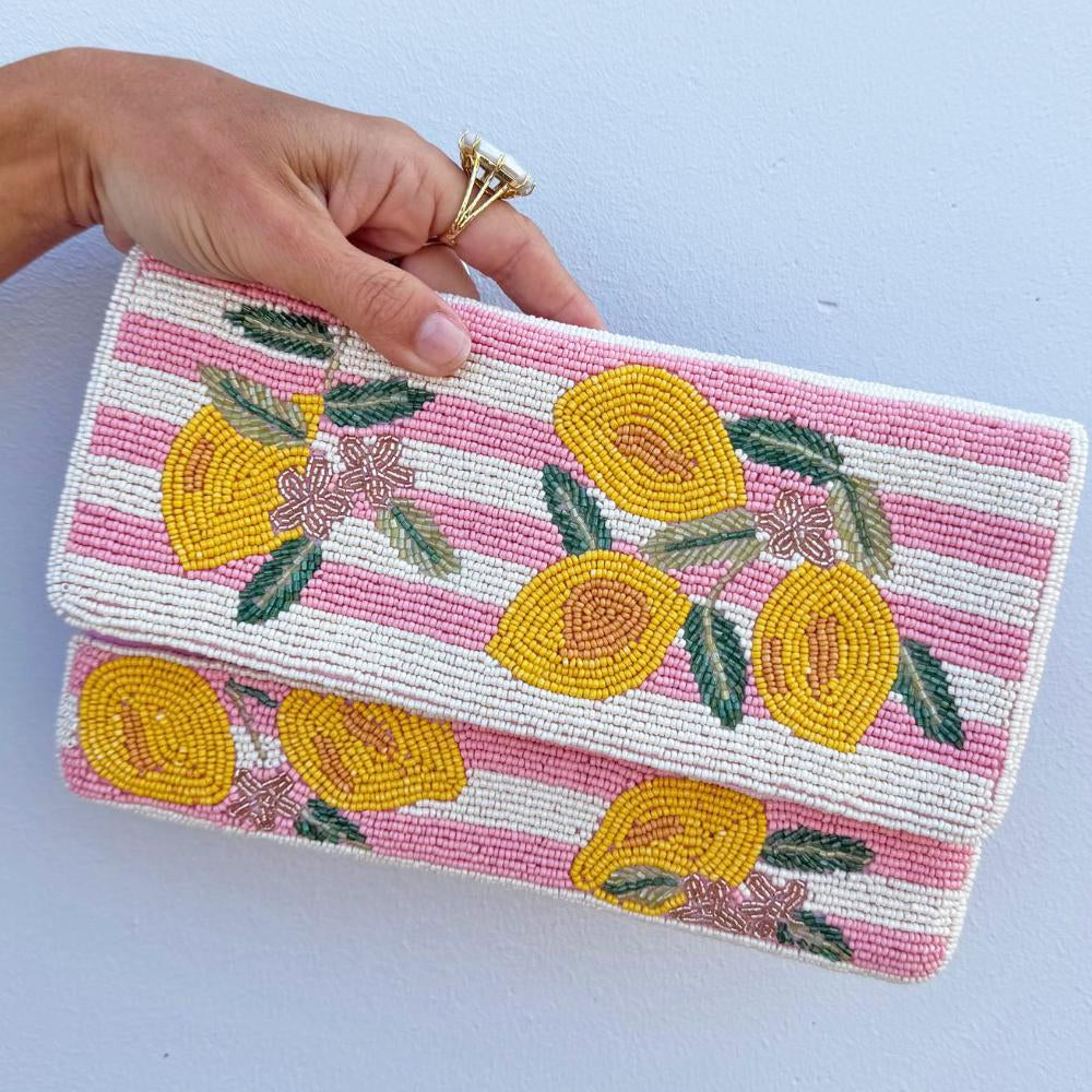 A hand holds a rectangular beaded clutch purse with a pink and white striped background and yellow lemon motifs, against a light blue wall.