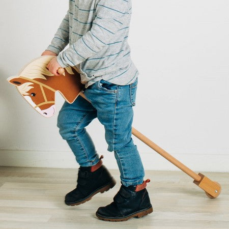 Bigjigs Toys Wooden Hobby Horse. Child holding a toy horse on a light wooden floor with a plain white wall background