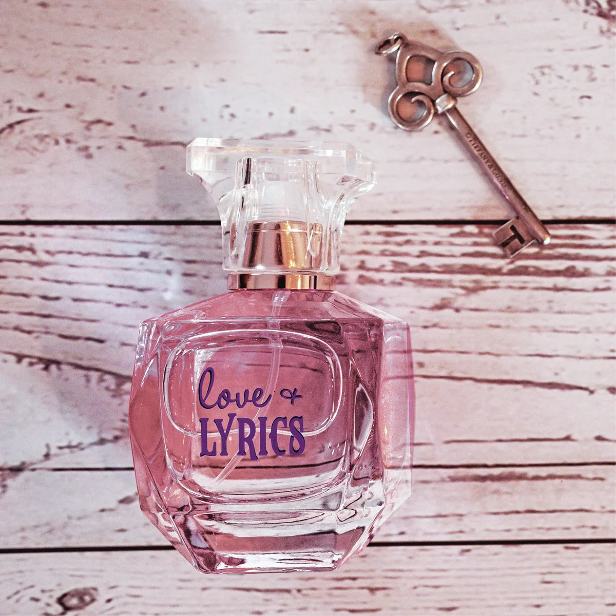 Pink perfume bottle labeled 'Love & Lyrics' on a wooden surface with a key.