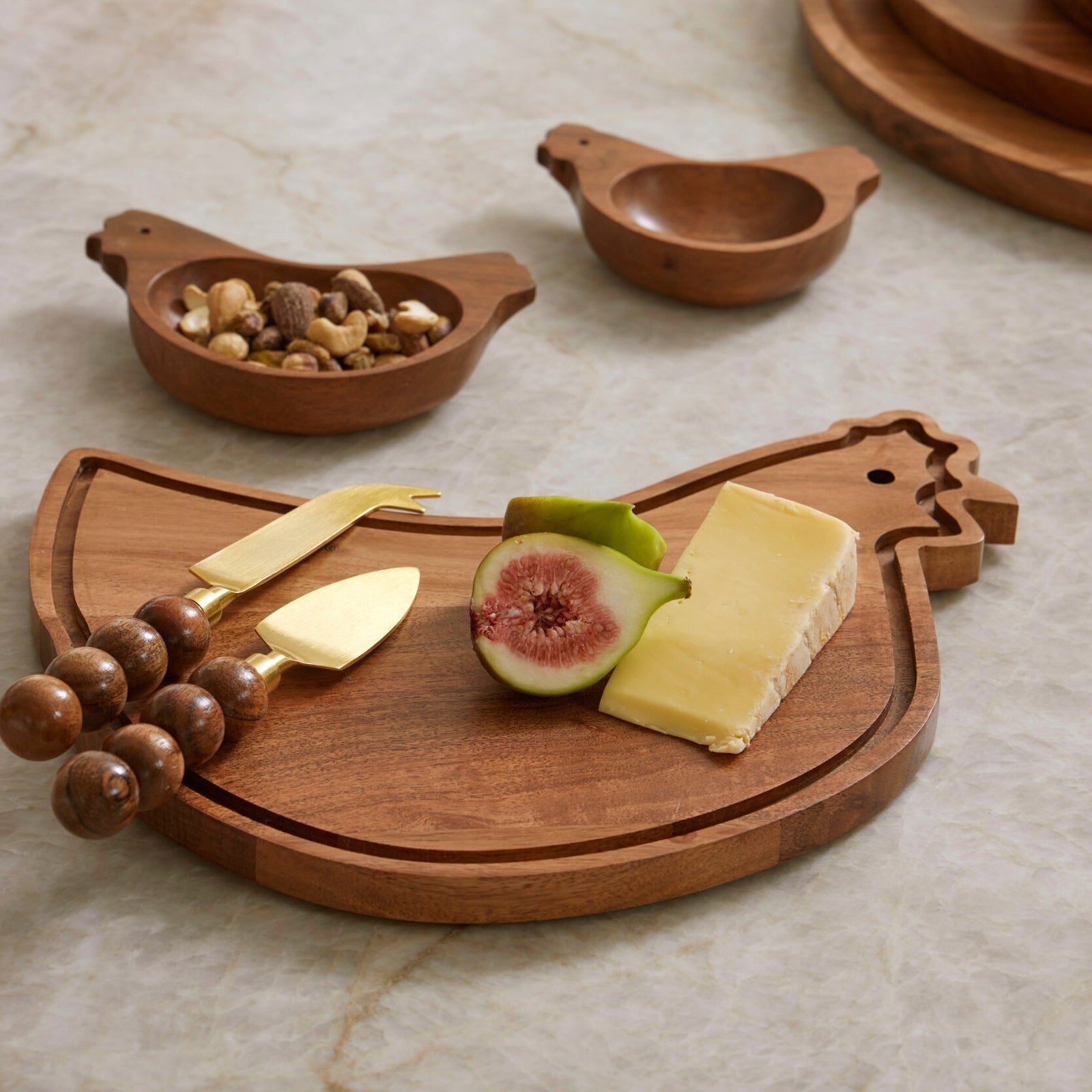 Wooden chicken-shaped cutting board with cheese, figs, and nuts on a marble surface.