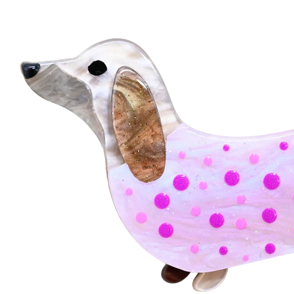 A dachshund-shaped decorative object with a pearlescent white and gray head, a brown marbled ear, and a pink body with purple polka dots and glitter, shown close up on a light background.