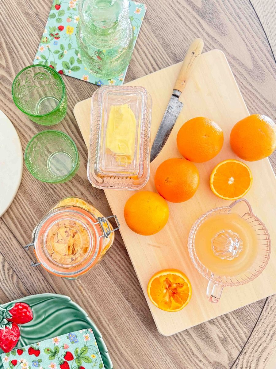 An overhead styled flat-lay of citrus ingredients and kitchen items on a wooden table, including whole and sliced oranges, a glass juicer, a glass bottle, a small jar, a butter dish with yellow butter, and a knife on a wooden cutting board.
