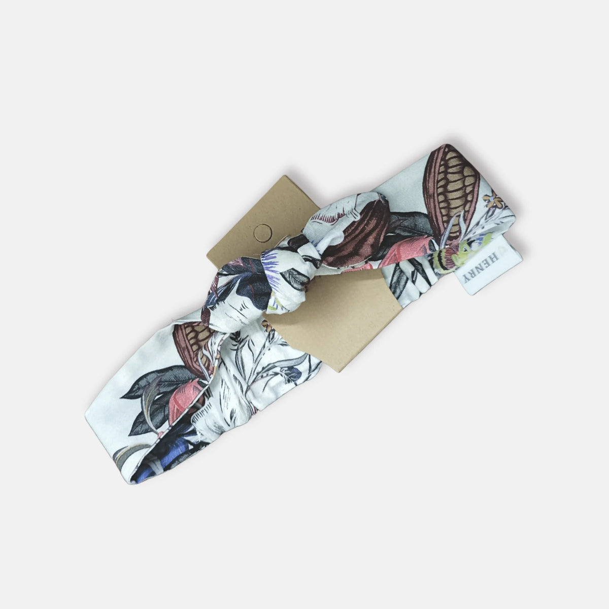 A knotted fabric headband with a vintage botanical illustration print in muted pink, brown, blue, and black on a white base, displayed on a brown kraft paper backing card against a light gray background.