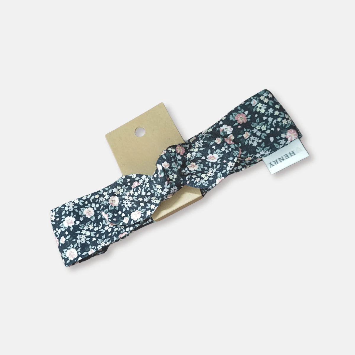 A knotted fabric headband with a dark navy blue floral pattern, featuring small pink, white, and green flowers. It is displayed on a light beige card with a small white brand tag that reads 'HENNY'.