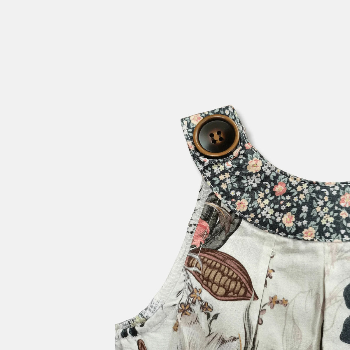 Close-up of a children's sleeveless top or dress featuring a dark floral collar with a large brown button and a light cream body with botanical and bird illustrations.