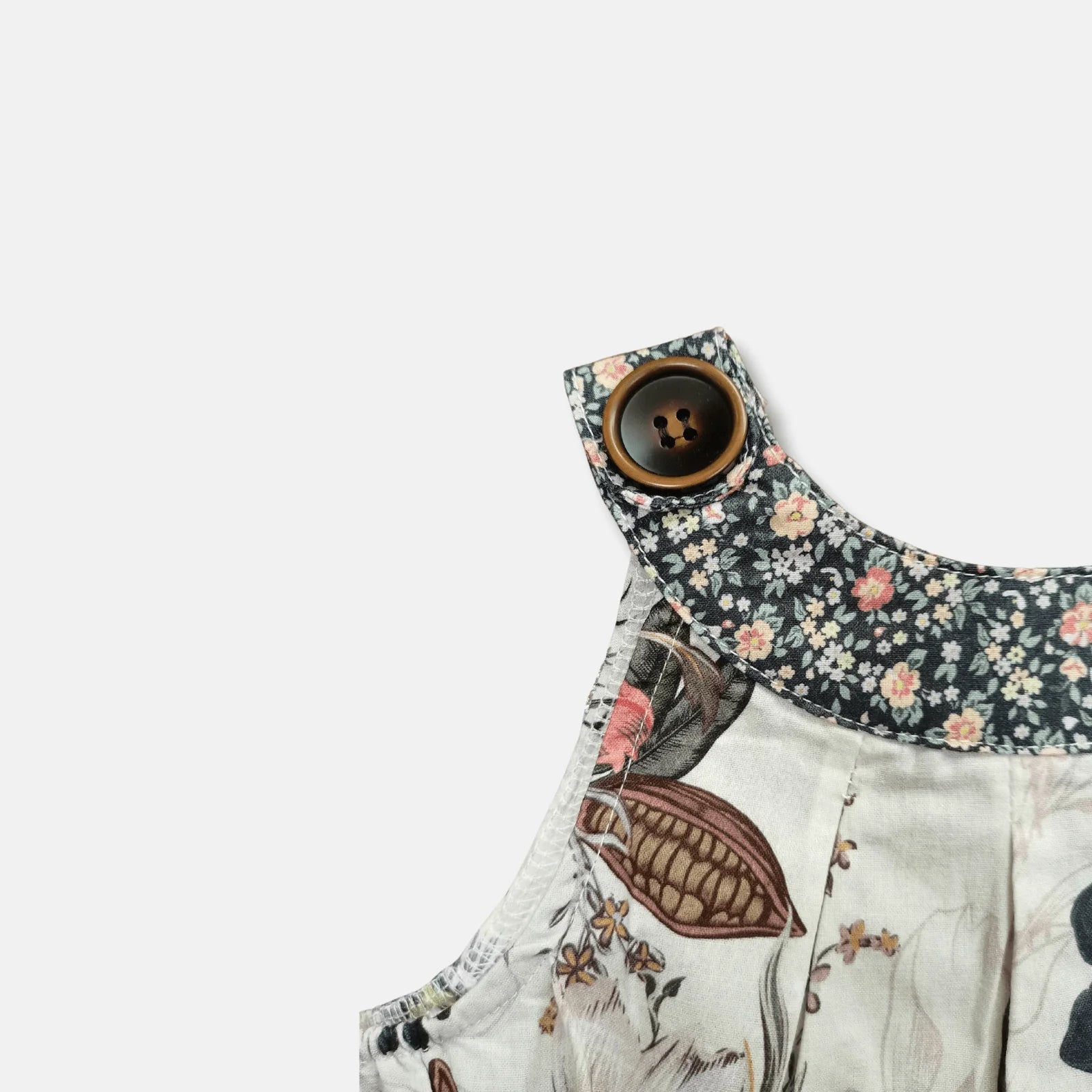Close-up of a children's sleeveless top or dress featuring a dark floral collar with a large brown button and a light cream body with botanical and bird illustrations.