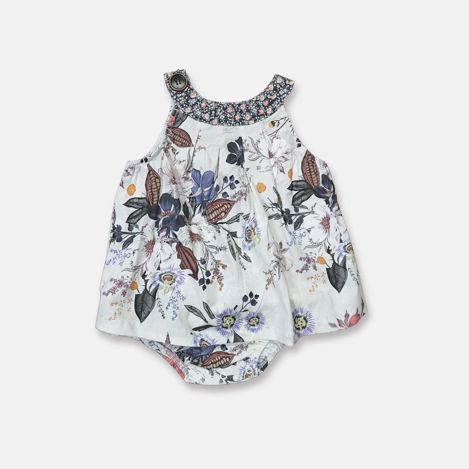 A toddler girl stands indoors wearing a sleeveless floral romper with a dark patterned collar and white sandals, photographed in a bright room with sheer curtains and sunlight on a light wood floor.