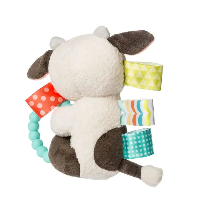 A plush cow baby toy shown from the back, featuring a cream-colored body, dark brown patches, a mint green bead handle, and colorful patterned fabric tabs in yellow, red, blue, and white.