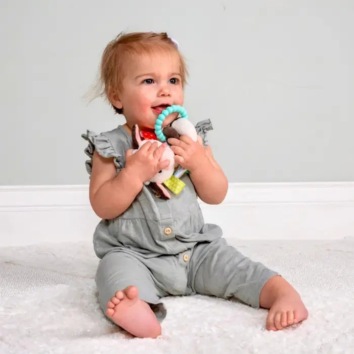 A baby sitting on a white textured rug holding a plush toy with a turquoise ring, wearing a light gray ruffle-shoulder romper.