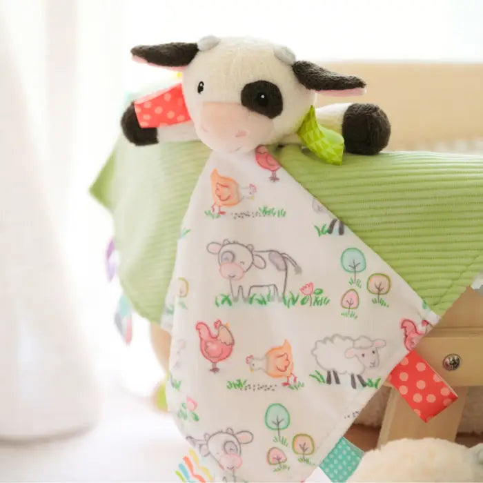 A soft baby lovey blanket featuring a plush cow with black ears and eyes, a green leaf accent, and a white fabric panel printed with cute farm animals like a cow, chicken, sheep, and trees. The blanket is draped over a light green ribbed baby crib or cushion.