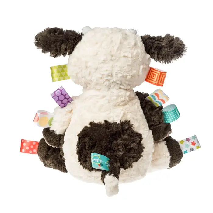 A plush cow sensory toy shown from the back, featuring a soft cream-colored body with dark brown patches, dark brown ears, and colorful patterned fabric tabs attached around it, photographed on a solid white background.