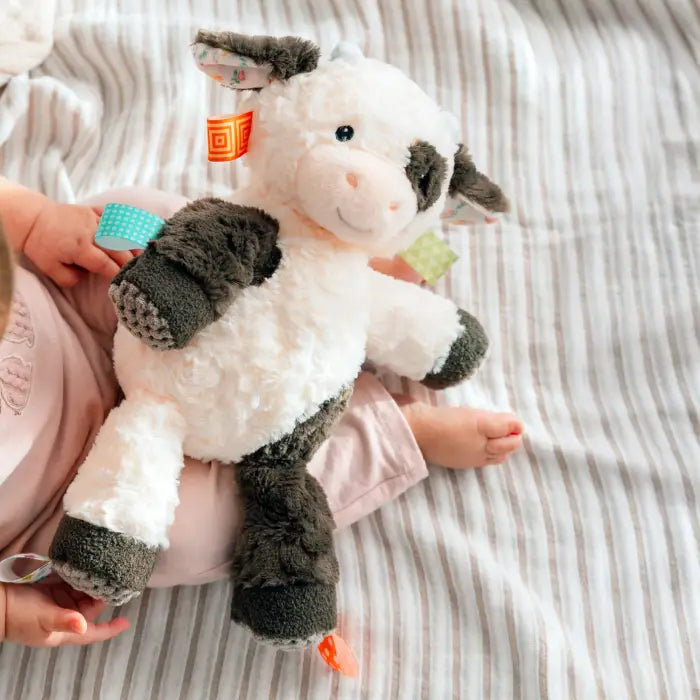 A plush cow toy being held by a baby on a striped bedsheet background. The cow is white with dark gray patches and has colorful fabric tags attached.
