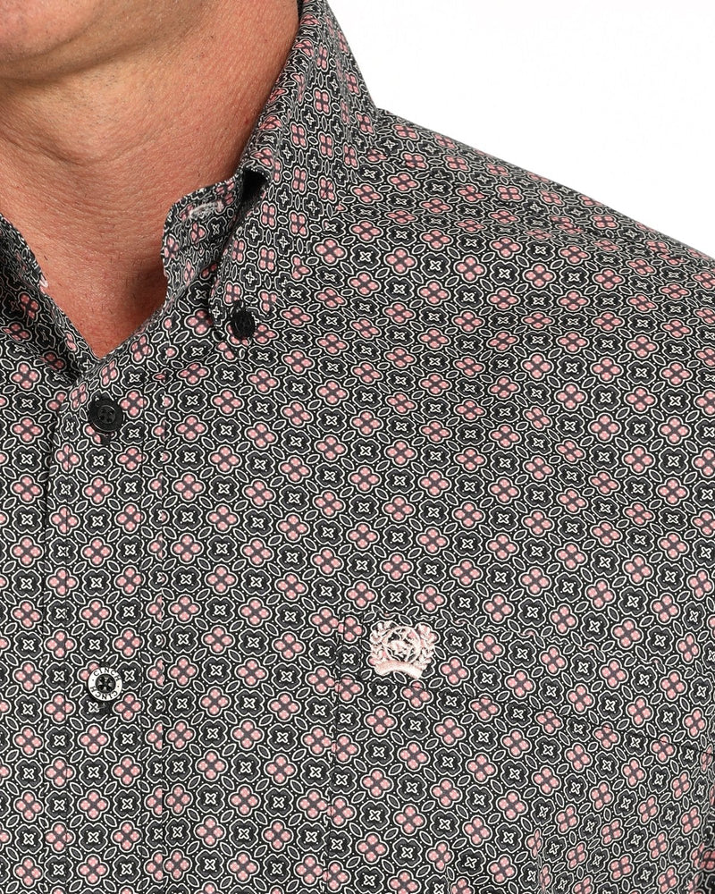 Close-up of a men's button-down shirt featuring a dense geometric pattern in black, pink, and white. The collar is open, showing the neck, and a small embroidered logo is visible on the chest.