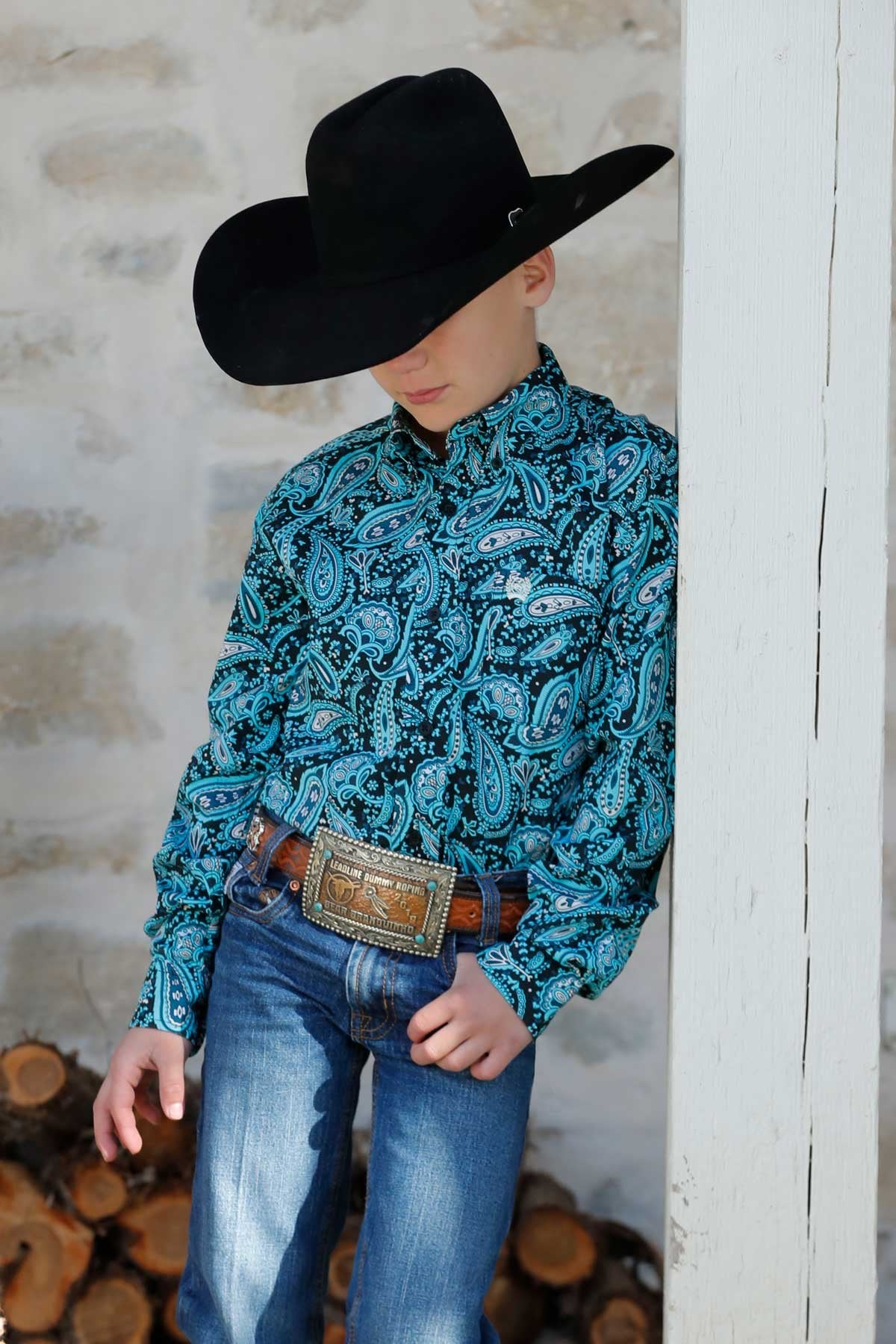 Cinch Boy's Paisley Print L/S Western Shirt. Person wearing a black cowboy hat and blue patterned shirt standing against a white wall with wooden logs below.