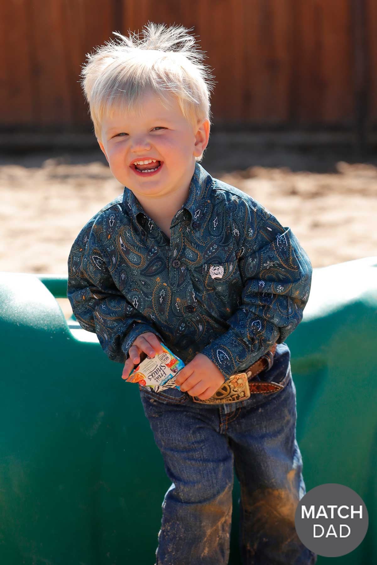 Cinch Paisley Print Toddler L/S Western Shirt. Child in a patterned shirt and jeans holding a toy, standing on a green surface with a wooden fence in the background.