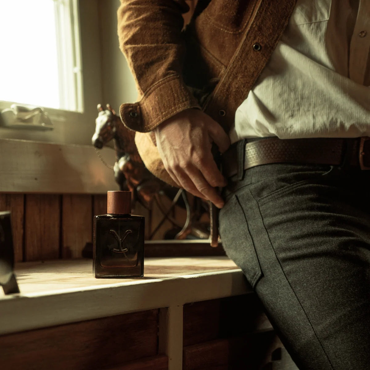 Person in a brown jacket and white shirt adjusting their belt with a bottle of cologne on a wooden surface.