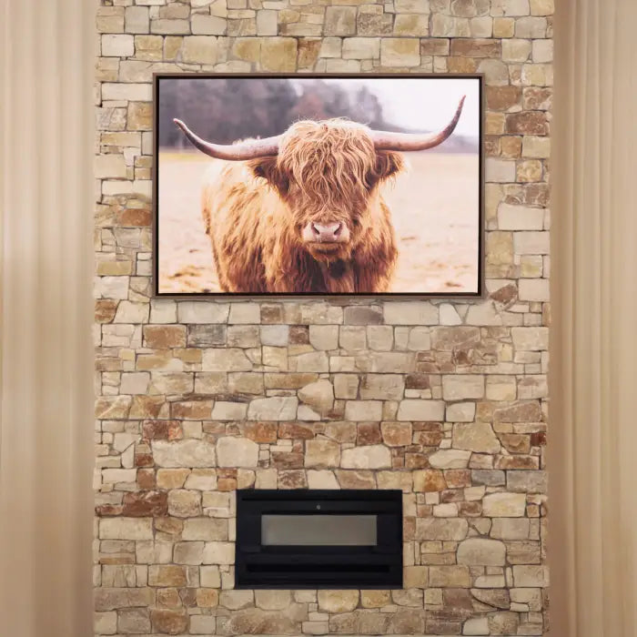A framed wall art print of a shaggy Highland cow mounted on a beige stone wall above a black fireplace insert.