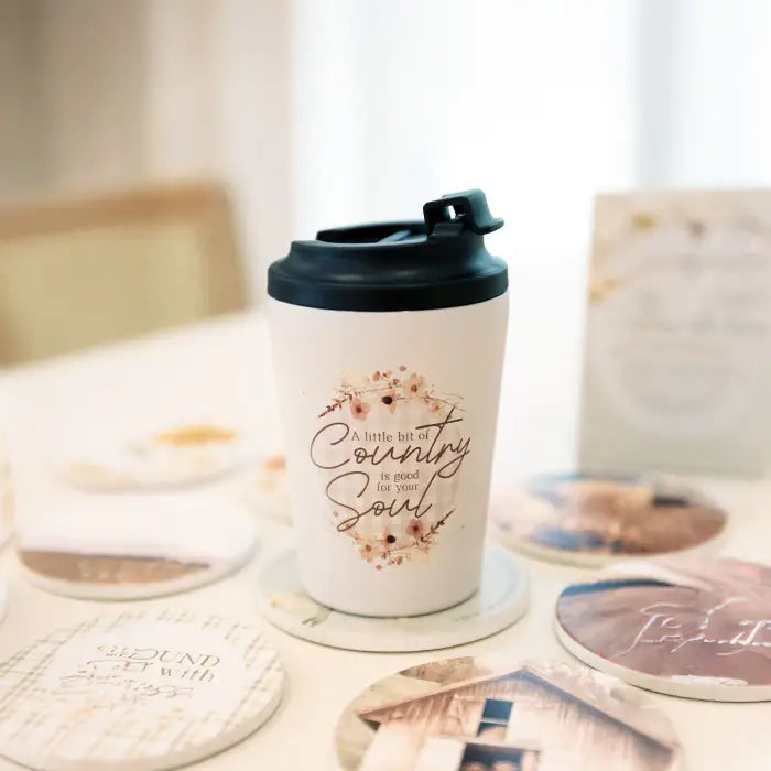 A white ceramic travel tumbler with a black lid sits on a table, printed with the phrase 'a little bit Country Soul' in brown script with a floral design. The background is softly blurred with other matching printed coasters and cards.