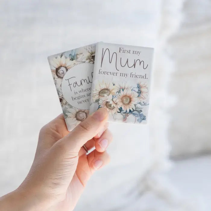 A hand holds two small square floral cards with printed quotes, one reading 'First my Mum forever my friend.' and the other partially visible reading 'Family is where it begins and never.