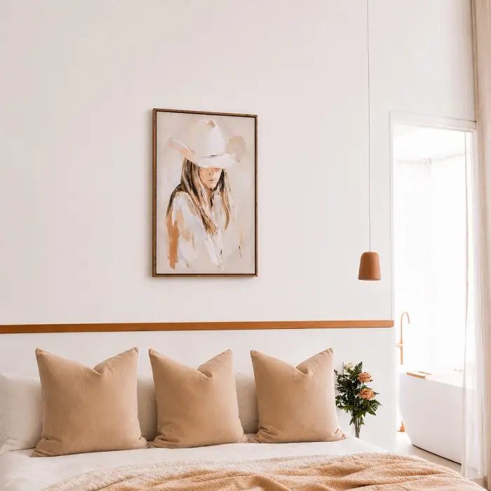 A framed art print of a woman in a cowboy hat hanging on a white wall above a bed with beige pillows and a beige blanket in a bright, modern bedroom.