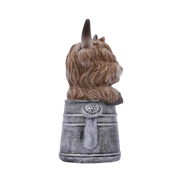 A decorative figurine of a shaggy brown Highland cow with dark horns, perched on top of a weathered gray metal bucket, shown against a plain white background.