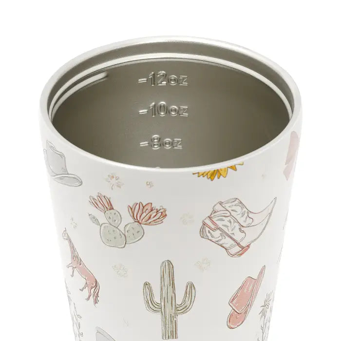 Close-up of a white insulated tumbler with a western-themed print featuring cacti, cowboy boots, hats, and a horse. The interior shows stainless steel lining with liquid measurement markings for 12oz, 10oz, and 8oz.