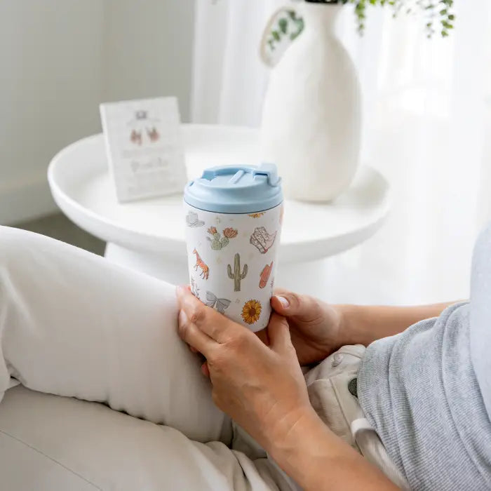A person sitting indoors holds a white reusable tumbler with a light blue lid. The tumbler is printed with small desert-themed illustrations including cacti, a sunflower, and a small animal.