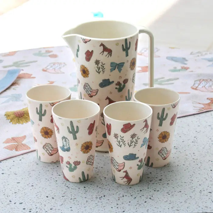 An off-white ceramic pitcher with a western-themed print featuring cowboy boots, cacti, sunflowers, horseshoes, and horses.