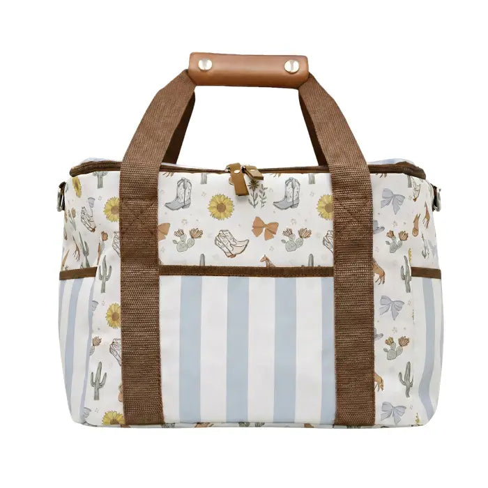 A canvas duffel bag with a western-themed print featuring cacti, cowboy boots, and butterflies, accented by brown webbed handles and a brown horizontal stripe.