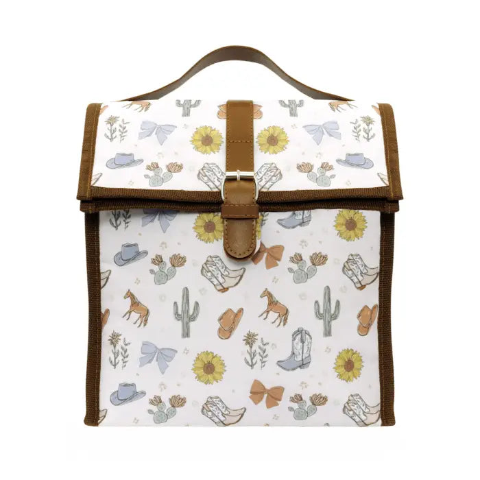 A canvas tote bag with a western-themed print featuring cowboy boots, cacti, sunflowers, and hats, finished with brown faux-leather trim and a top handle.