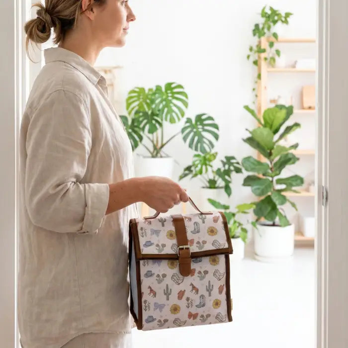 A woman in a light beige shirt holds a small rectangular picnic-style bag with a white background and a colorful cactus and floral print, featuring brown trim and a top handle, in a bright doorway with potted plants in the background.