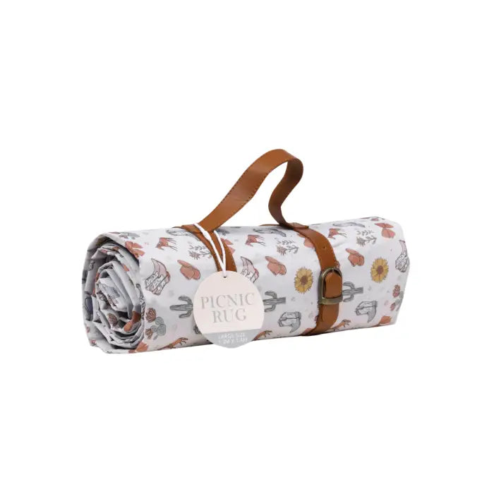 A rolled-up picnic rug with a white background and a multicolor western-themed print, featuring brown faux-leather straps and a hanging tag that reads 'PICNIC RUG'.