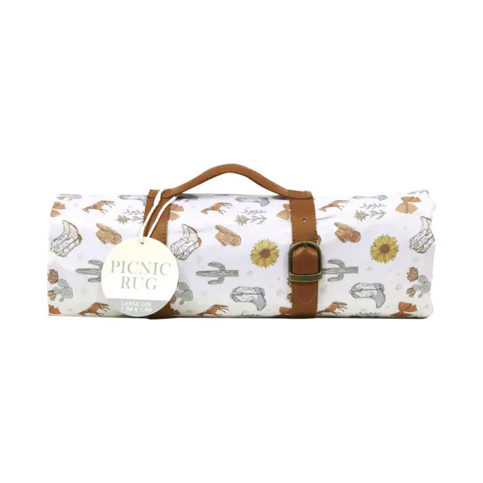 A rolled-up picnic rug with a white background and a desert-themed print of cacti, sunflowers, and desert animals. It features a brown leather handle and a matching brown leather strap with a metal buckle, along with a round paper tag that reads 'PICNIC RUG'.