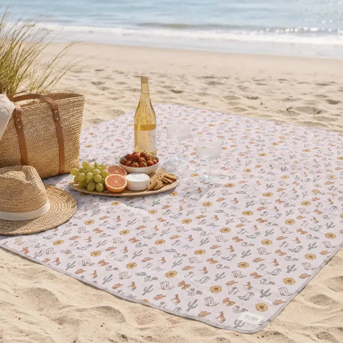 A square picnic blanket with a white background and a small cactus and floral pattern is spread out on a sandy beach. A straw hat, woven straw tote bag, and a bottle of white wine are arranged on the blanket along with grapes, a sliced orange, a small bowl of nuts, and two clear drinking glasses.