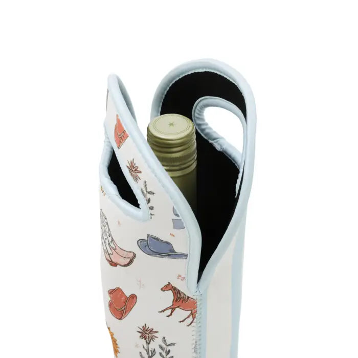 A close-up of a white neoprene wine bottle holder with a light blue trim and handle, featuring a western-themed print of cowboy hats, boots, and a horse. A wine bottle is partially inserted into the holder.