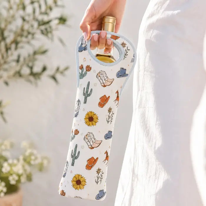 A hand holds a wine bottle in a white wine bottle sleeve with a western-themed print of cowboy boots, cacti, sunflowers, and hats. The sleeve has a light blue trim and is shown against a soft, bright background with a person in a white dress partially visible.