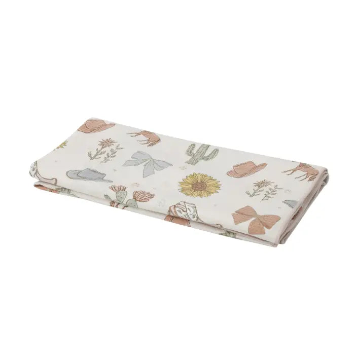 A folded fabric item, likely a blanket or quilt, featuring a western-themed print with a sunflower, cactus, boots, and bows on a cream background.